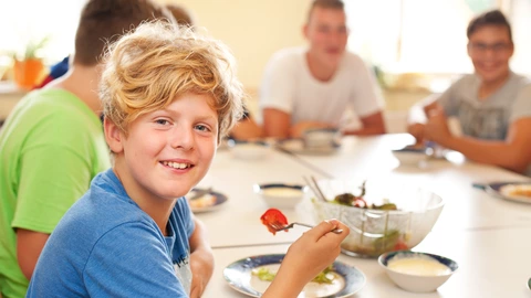 Ein Junge isst Salat. Im Hintergrund sitzen mehrere Personen am Tisch.