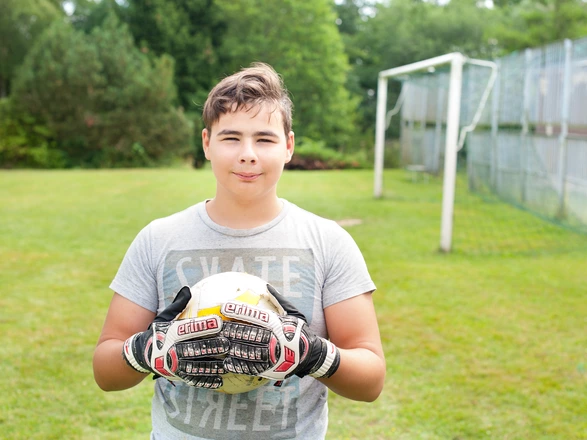 Ein Junge steht auf dem Rasen und hält einen Fußball in der Hand. Im Hintergrund ist ein Fußballtor.