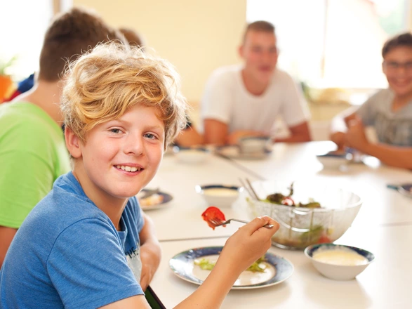 Ein Junge isst Salat. Im Hintergrund sitzen mehrere Personen am Tisch.