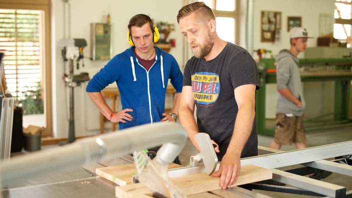 Ein Ausbilder und ein Jugendlicher arbeiten mit Holzbrettern an einer Maschine.